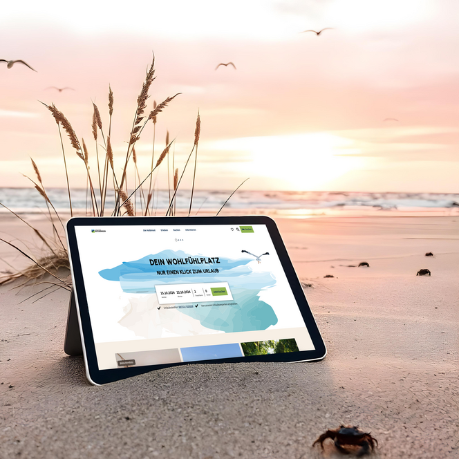 Ein Tablet mit geöffneter Website von Butjadingen steht am Sandstrand bei Sonnenuntergang, während Möwen fliegen und vorn eine kleine Krabbe zu sehen ist.