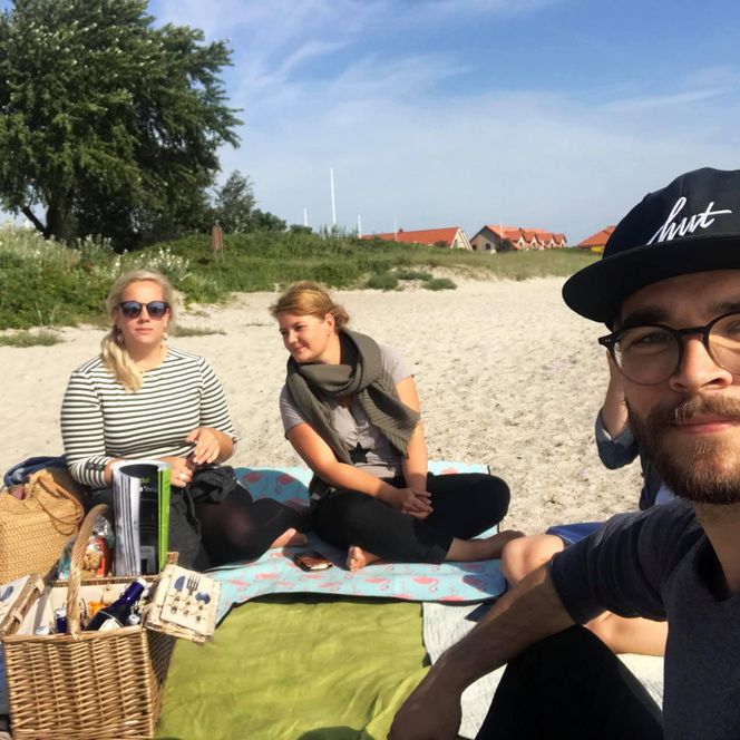Drei junge Erwachsene sitzen auf Picknickdecken am Sandstrand, ein Picknickkorb steht vor ihnen, im Hintergrund sind grüne Büsche, Bäume und Häuser mit roten Dächern zu sehen