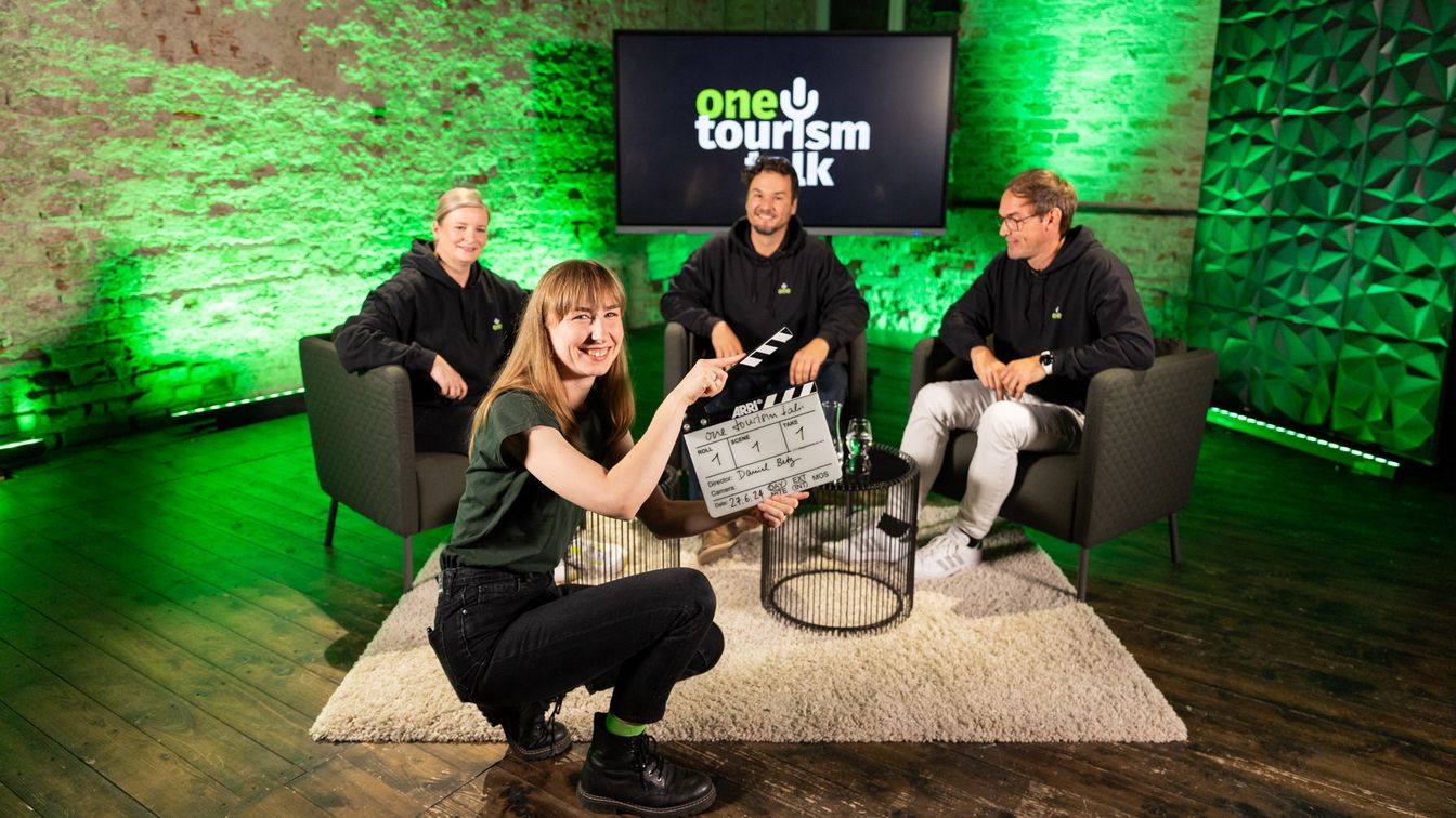 Drei Talkgäste sitzen auf einer grün beleuchteten Bühne mit dem Schriftzug "one tourism.talk" im Hintergrund. Davor hockt eine junge Frau mit einer Regie-Klappe