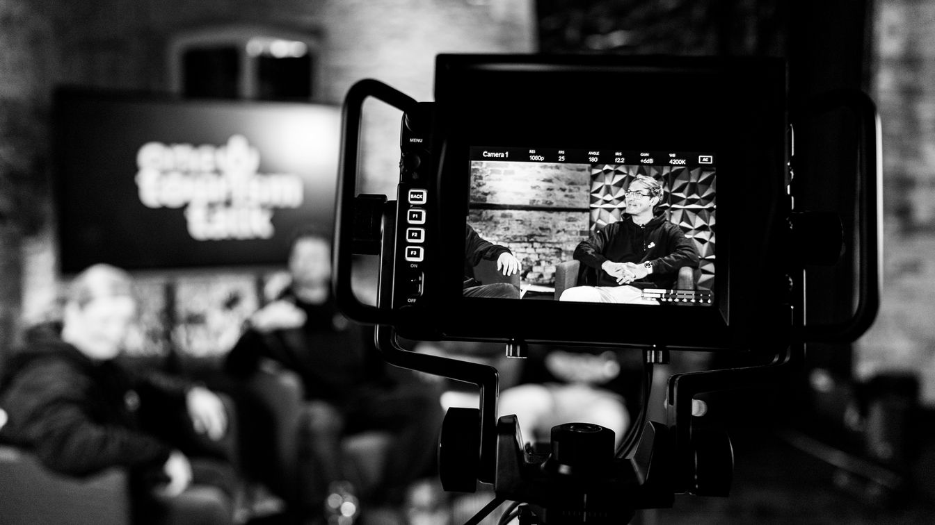 Schwarz-weiß-Foto einer Talkshow-Aufnahme, bei der der Kamerabildschirm zwei Personen im Gespräch auf einem Set zeigt