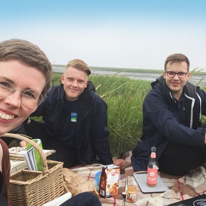 Drei junge Menschen sitzen lachend bei einem Picknick auf einer Decke im Gras, im Hintergrund ist eine weite Landschaft mit Wasser und Himmel zu sehen