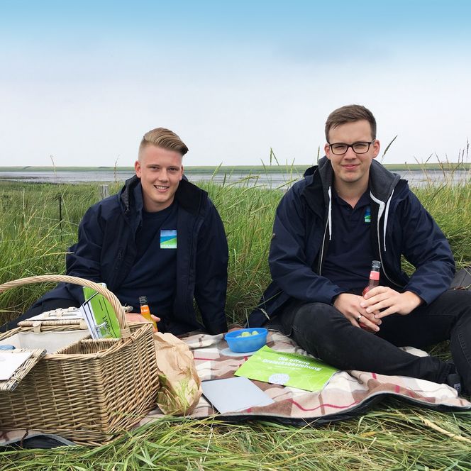 Zwei junge Männer sitzen auf einer Picknickdecke im Gras an einem Gewässer, haben Picknickutensilien und Getränke vor sich und lächeln in die Kamera