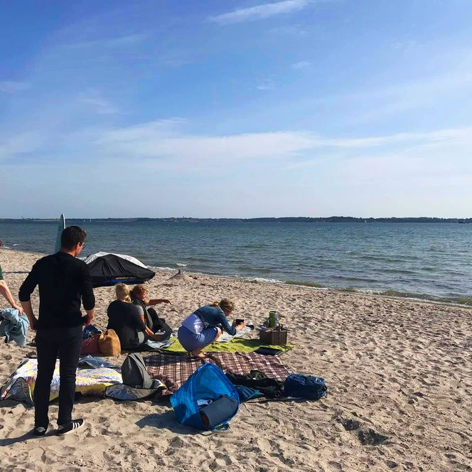 Mehrere Personen sitzen und stehen bei einem Picknick am Sandstrand, mit Decken, Taschen und Picknickkorb, während im Hintergrund das Meer und ein weiter Himmel zu sehen sind