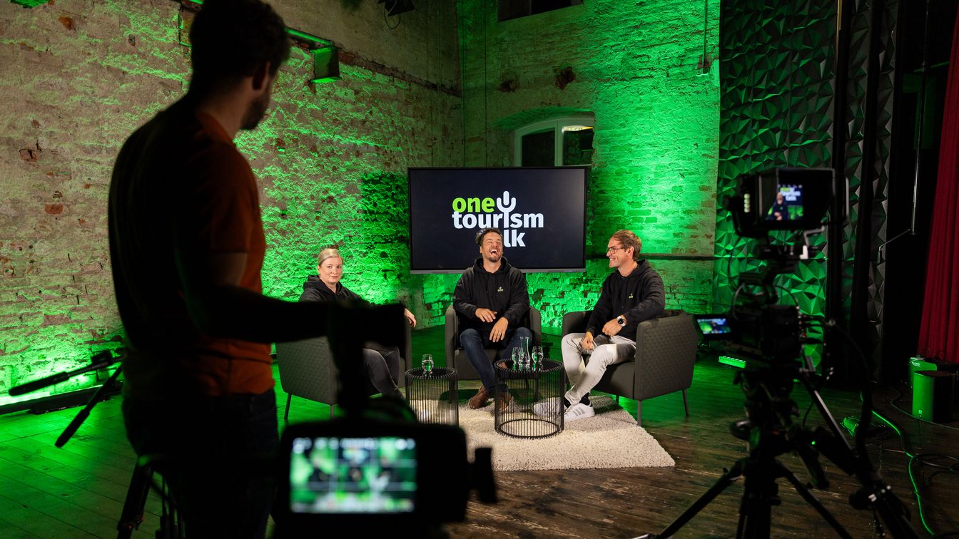 Drei Personen sitzen auf einem Talkshow-Set mit grüner Beleuchtung, während Kameras und ein Kameramann im Vordergrund sichtbar sind.
