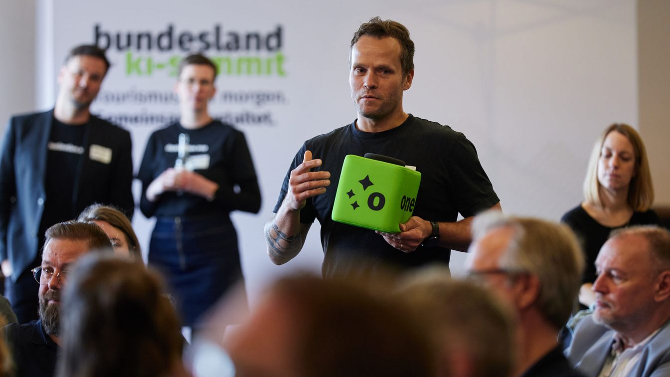 Freie Wortmeldung beim Bundesland KI‑Summit vor Publikum mit einem grünen Mikrofonwürfel mit one‑Branding
