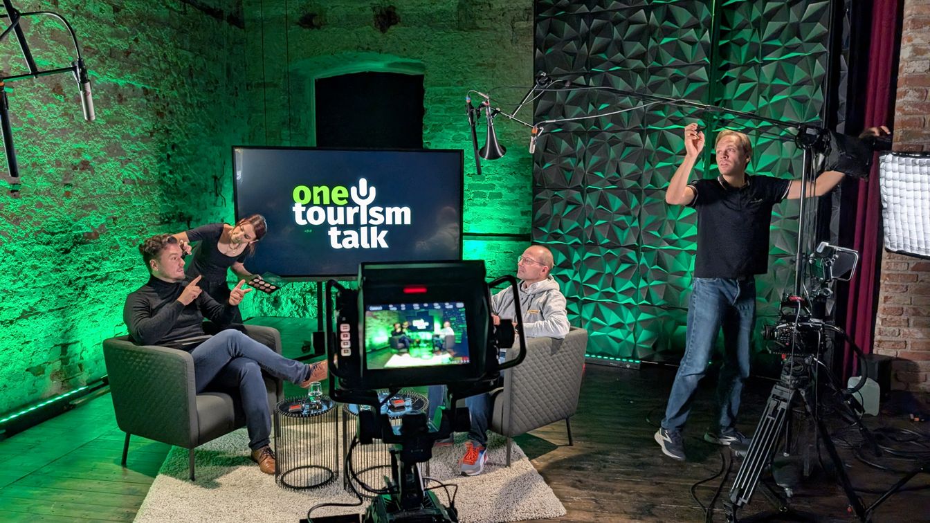 Zwei Männer sitzen auf einem Talkshow-Set, während eine Visagistin einen Gast schminkt und ein Techniker das Equipment vorbereitet; im Hintergrund leuchtet grüne Beleuchtung.